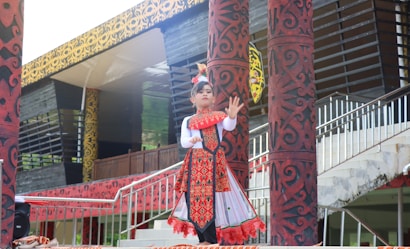 A child is dressed in traditional clothing, performing a cultural dance. The outfit is decorated with intricate patterns and vibrant colors, predominantly red and white. The background includes ornately designed pillars and a building with a patterned facade.