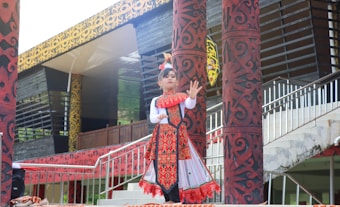 A child is dressed in traditional clothing, performing a cultural dance. The outfit is decorated with intricate patterns and vibrant colors, predominantly red and white. The background includes ornately designed pillars and a building with a patterned facade.