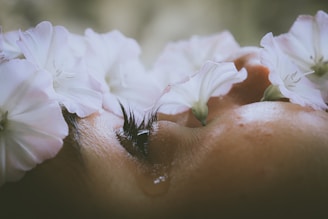 A close-up of a person's face, partially obscured by delicate white flowers. The eyelashes and a single tear are visible, conveying a sense of emotion.