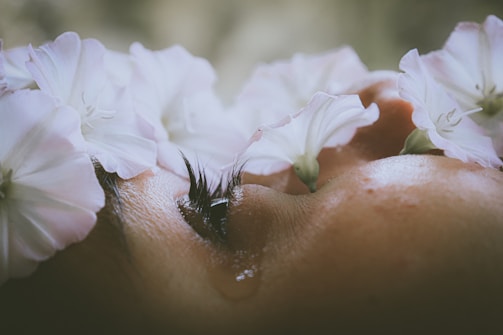 A close-up of a person's face, partially obscured by delicate white flowers. The eyelashes and a single tear are visible, conveying a sense of emotion.
