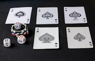 Five playing cards, all aces of spades, are arranged on a dark surface. Beside the cards, there is a stack of poker chips with two dice on top. Three additional dice are placed nearby, with both the poker chips and dice featuring a combination of red, blue, and white colors.