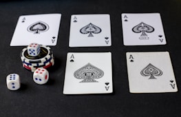 Five playing cards, all aces of spades, are arranged on a dark surface. Beside the cards, there is a stack of poker chips with two dice on top. Three additional dice are placed nearby, with both the poker chips and dice featuring a combination of red, blue, and white colors.