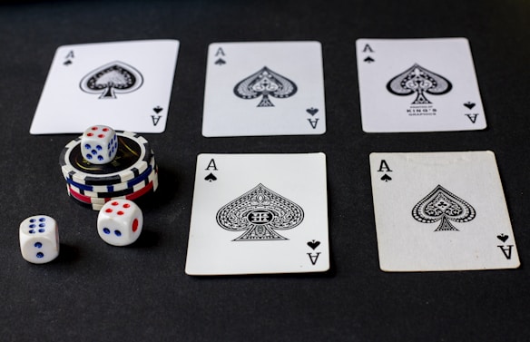 Five playing cards, all aces of spades, are arranged on a dark surface. Beside the cards, there is a stack of poker chips with two dice on top. Three additional dice are placed nearby, with both the poker chips and dice featuring a combination of red, blue, and white colors.