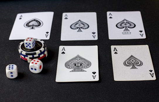 Five playing cards, all aces of spades, are arranged on a dark surface. Beside the cards, there is a stack of poker chips with two dice on top. Three additional dice are placed nearby, with both the poker chips and dice featuring a combination of red, blue, and white colors.