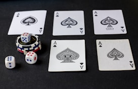 Five playing cards, all aces of spades, are arranged on a dark surface. Beside the cards, there is a stack of poker chips with two dice on top. Three additional dice are placed nearby, with both the poker chips and dice featuring a combination of red, blue, and white colors.