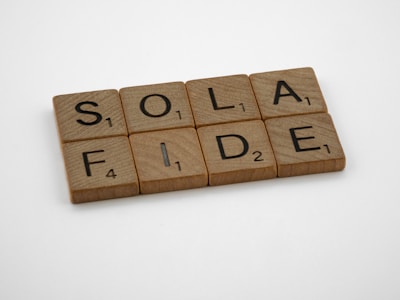 Wooden Scrabble tiles arranged to spell 'SOLA FIDE' on a plain white background. Each tile features a letter with its corresponding point value in black print.