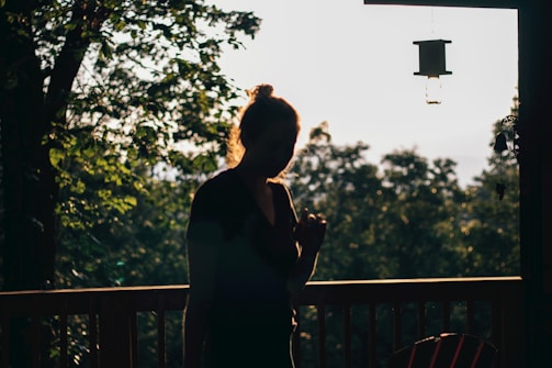 A person is silhouetted against a sunlit forest backdrop, with lush green leaves and wooden railings. A bird feeder hangs nearby, casting soft shadows.
