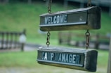 Two wooden signs hanging in a row, both with dark green and white text. The top sign reads 'WELCOME' and the bottom sign reads 'CAMP MANAGER'. The signs appear aged, with a rustic, outdoor look, suspended by metal chains against a blurry green background.