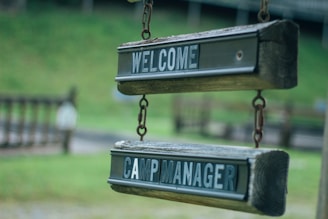 Two wooden signs hanging in a row, both with dark green and white text. The top sign reads 'WELCOME' and the bottom sign reads 'CAMP MANAGER'. The signs appear aged, with a rustic, outdoor look, suspended by metal chains against a blurry green background.