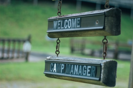Two wooden signs hanging in a row, both with dark green and white text. The top sign reads 'WELCOME' and the bottom sign reads 'CAMP MANAGER'. The signs appear aged, with a rustic, outdoor look, suspended by metal chains against a blurry green background.
