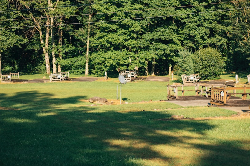 A lush green landscape featuring multiple wooden picnic tables and benches scattered across a grassy area. The background consists of dense, leafy trees, casting shadows on the ground, while a clear sky is visible above. A satellite dish and a few small decorative rocks are also present in the scene.