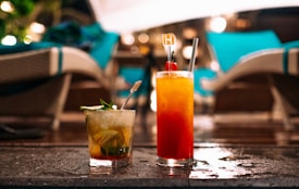 Two elegant cocktails are placed on a wet surface, with lounge chairs and soft ambient lighting in the background, suggesting a relaxed, evening setting. One drink is vibrant red with a cherry garnish, and the other is a minty, citrusy cocktail, each in a tall glass.