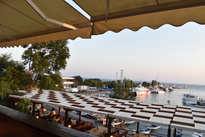 A dining area with people sitting under a decorative awning overlooks a scenic harbor filled with boats. A tree and some greenery are nearby, while the calm water and boats create a peaceful atmosphere. In the distance, buildings and hills can be seen under a clear sky.