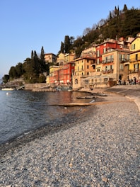 Charming lakeside village with colorful buildings perched along the shore. The architecture features vibrant facades in hues of red, yellow, and orange. The calm lake reflects the warm tones of the buildings and the sky, creating a picturesque setting. A pebble beach stretches along the water's edge, leading to a small group of people enjoying the scenic view. Lush greenery with tall trees surrounds the area, adding to the serene atmosphere.