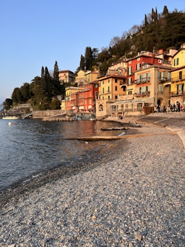 Charming lakeside village with colorful buildings perched along the shore. The architecture features vibrant facades in hues of red, yellow, and orange. The calm lake reflects the warm tones of the buildings and the sky, creating a picturesque setting. A pebble beach stretches along the water's edge, leading to a small group of people enjoying the scenic view. Lush greenery with tall trees surrounds the area, adding to the serene atmosphere.