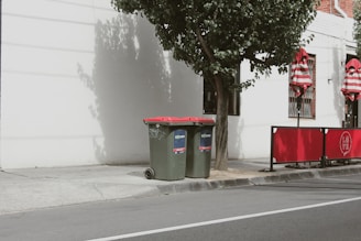 Two green garbage bins with red lids stand next to a tree on a sidewalk. The backdrop features a white building wall with shadows cast on it, alongside red and white striped umbrellas behind a red barrier fence.