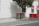 Two green garbage bins with red lids stand next to a tree on a sidewalk. The backdrop features a white building wall with shadows cast on it, alongside red and white striped umbrellas behind a red barrier fence.