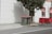 Two green garbage bins with red lids stand next to a tree on a sidewalk. The backdrop features a white building wall with shadows cast on it, alongside red and white striped umbrellas behind a red barrier fence.