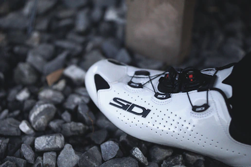 A white cycling shoe with black accents and a sleek design lies on a bed of gray stones. The shoe features a prominent brand logo and a sophisticated lacing system.