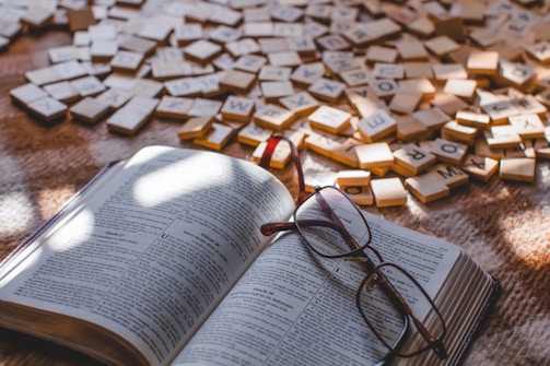 An open book is placed on a surface with reading glasses resting on it. Scattered around are numerous wooden letter tiles, creating a casual and intellectual atmosphere.
