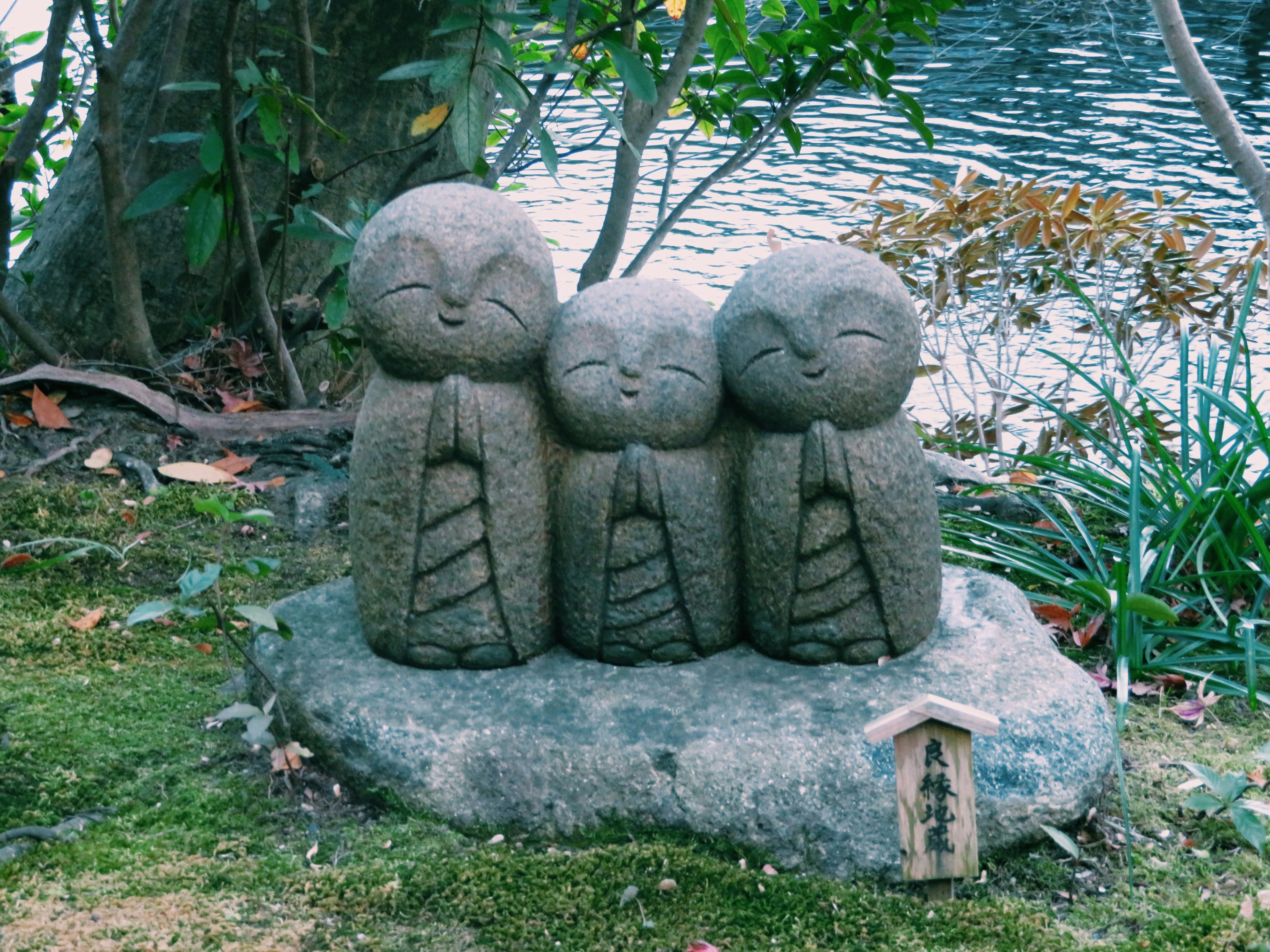 Three stone Jizo statues sit on a moss-covered rock beside a calm pond, framed by garden foliage.
