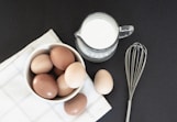 a bowl of eggs next to a whisk and a glass of milk