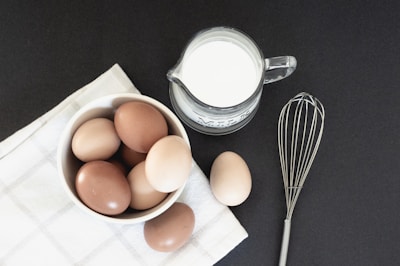 A collection of brown and tan eggs is placed in a white bowl atop a white checkered cloth. Next to the bowl, there is a glass pitcher filled with milk, labeled with the word 'milk.' A metal whisk is positioned nearby, set against a dark background.