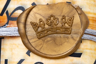 A gold wax seal embossed with a crown emblem is placed over a textured background featuring intertwined white threads. The background includes a mix of muted and warm colors with partial black text visible.