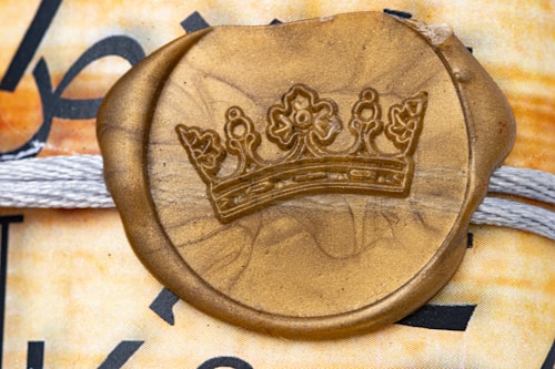 A gold wax seal embossed with a crown emblem is placed over a textured background featuring intertwined white threads. The background includes a mix of muted and warm colors with partial black text visible.