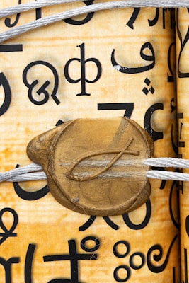 A close-up image of a scroll with various black symbol characters printed on it. The scroll is tied with a white string and features a gold wax seal with a fish symbol embossed on it.