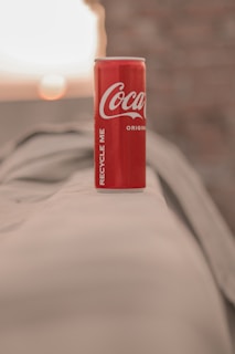 A red soda can with the brand logo is prominently displayed on a beige surface, with a blurred background featuring soft, warm lighting and hints of a brick wall.