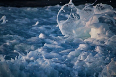 White sea foam splashes dynamically in mid-air above turbulent waves. The texture is vivid, with frothy patterns weaving across the surface. The light captures the foam, highlighting its movement and energy.