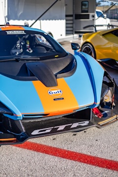 A race car with a striking orange and blue livery featuring the Gulf logo is parked next to another yellow car. The focus is on the front part of the car, showcasing its aerodynamic design and various sponsor logos. The setting appears to be a racetrack or a car event, with a trailer in the background.