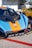 A race car with a striking orange and blue livery featuring the Gulf logo is parked next to another yellow car. The focus is on the front part of the car, showcasing its aerodynamic design and various sponsor logos. The setting appears to be a racetrack or a car event, with a trailer in the background.