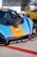 A race car with a striking orange and blue livery featuring the Gulf logo is parked next to another yellow car. The focus is on the front part of the car, showcasing its aerodynamic design and various sponsor logos. The setting appears to be a racetrack or a car event, with a trailer in the background.