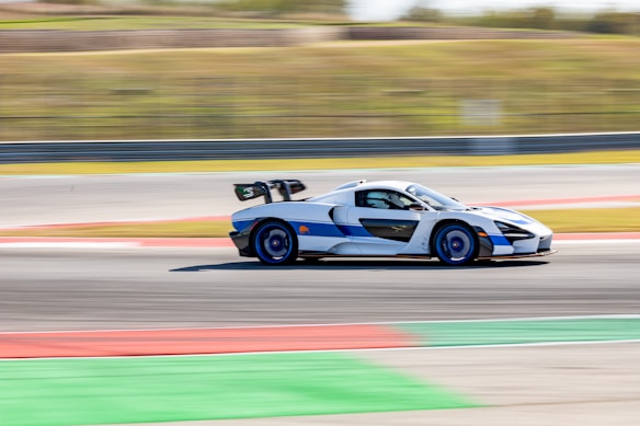 A high-performance sports car is captured in motion on a racing track. The car has a sleek aerodynamic design with a prominent rear wing and is predominantly silver and black with blue accents. The background is blurred, emphasizing the car's speed.