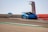 A bright blue sports car is racing on a track at high speed, with another similar car visible in the background. The track has a smooth surface with red and green markings, and a distinctive observation tower is in the background against a clear sky.