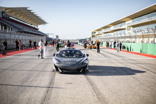 A sleek, gray safety car is positioned in the center of a race track, surrounded by people and various sports cars in the background. The scene takes place on a clear day with modern grandstands and safety barriers visible on either side of the track.