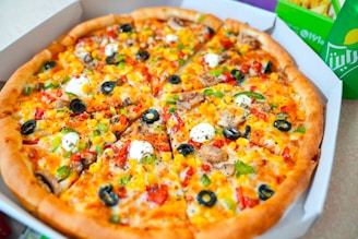 A freshly baked pizza topped with a variety of colorful ingredients, including slices of black olives, green and red bell peppers, mushrooms, corn, and dollops of white cheese or sauce. The pizza appears to be cut into slices and is served in a cardboard box. In the background, a green beverage can and fries are partially visible, suggesting a complete meal.