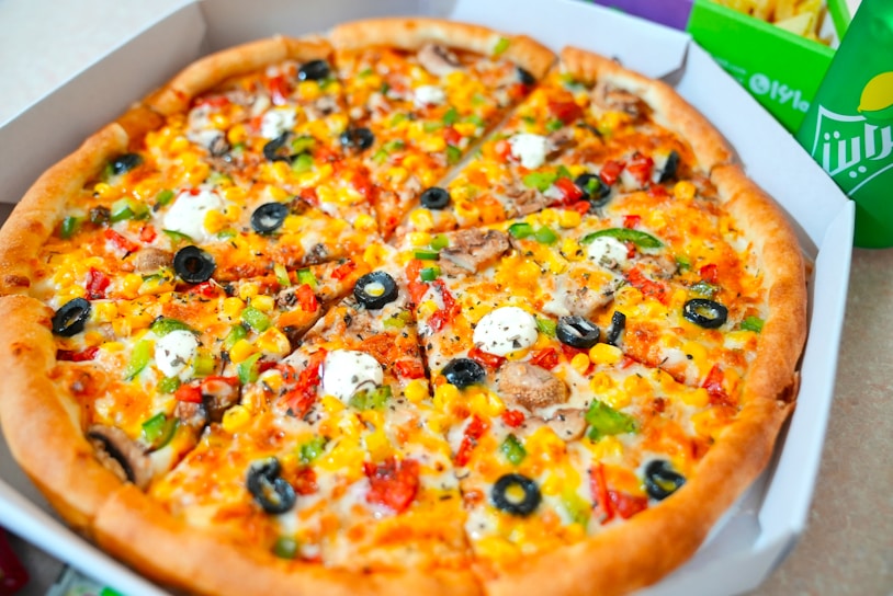 A freshly baked pizza topped with a variety of colorful ingredients, including slices of black olives, green and red bell peppers, mushrooms, corn, and dollops of white cheese or sauce. The pizza appears to be cut into slices and is served in a cardboard box. In the background, a green beverage can and fries are partially visible, suggesting a complete meal.