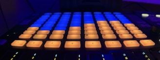 A close-up view of a music production controller with illuminated pads. The pads are lit in shades of orange and blue, arranged in a grid pattern on a dark background.