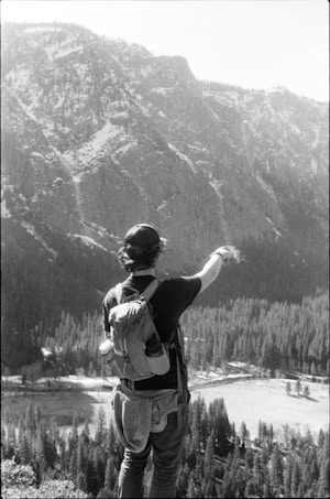 A person with a backpack stands on an elevated vantage point, pointing toward a vast landscape of a mountainous terrain. The scene is in black and white, adding a classic and nostalgic feel. The background shows rugged mountains and a dense forest below.