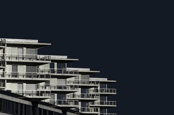 A modern, multi-story residential building with geometric patterns in its architecture. The structure features numerous balconies and large windows, with a stark contrast against a dark, uncluttered sky.