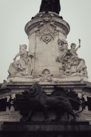 A tall, ornate stone monument adorned with intricate carvings and sculptures, featuring symbolic figures seated at its base. A prominent lion statue is positioned in front, conveying strength and majesty. French inscriptions are visible on the monument's surface, highlighting its cultural significance.
