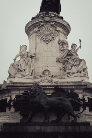 A tall, ornate stone monument adorned with intricate carvings and sculptures, featuring symbolic figures seated at its base. A prominent lion statue is positioned in front, conveying strength and majesty. French inscriptions are visible on the monument's surface, highlighting its cultural significance.