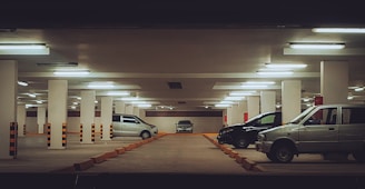 A dimly lit underground parking garage with multiple parked cars, including a silver van and a black sedan. The garage is supported by white pillars and has fluorescent lights on the ceiling. There are orange and black striped barriers adjacent to the parking spaces.