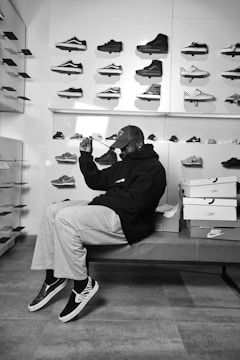 A person sitting on a bench in a sneaker store surrounded by a display of various sneakers on the wall. The individual is wearing casual clothing including a cap and hooded sweatshirt, sitting beside stacked shoe boxes.