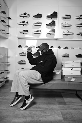 A person sitting on a bench in a sneaker store surrounded by a display of various sneakers on the wall. The individual is wearing casual clothing including a cap and hooded sweatshirt, sitting beside stacked shoe boxes.