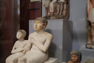 A collection of ancient sculptures depicted in a museum setting, featuring seated figures made from stone with remnants of paint. The figures exhibit calm, serene expressions and are placed on pedestals with other artifacts nearby.