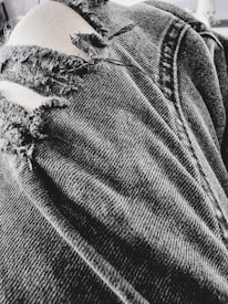 A close-up view of a pair of black, ripped denim jeans with several frayed tears exposing light-colored fabric beneath. The texture of the denim fabric is prominent, showing detailed stitching and wear.